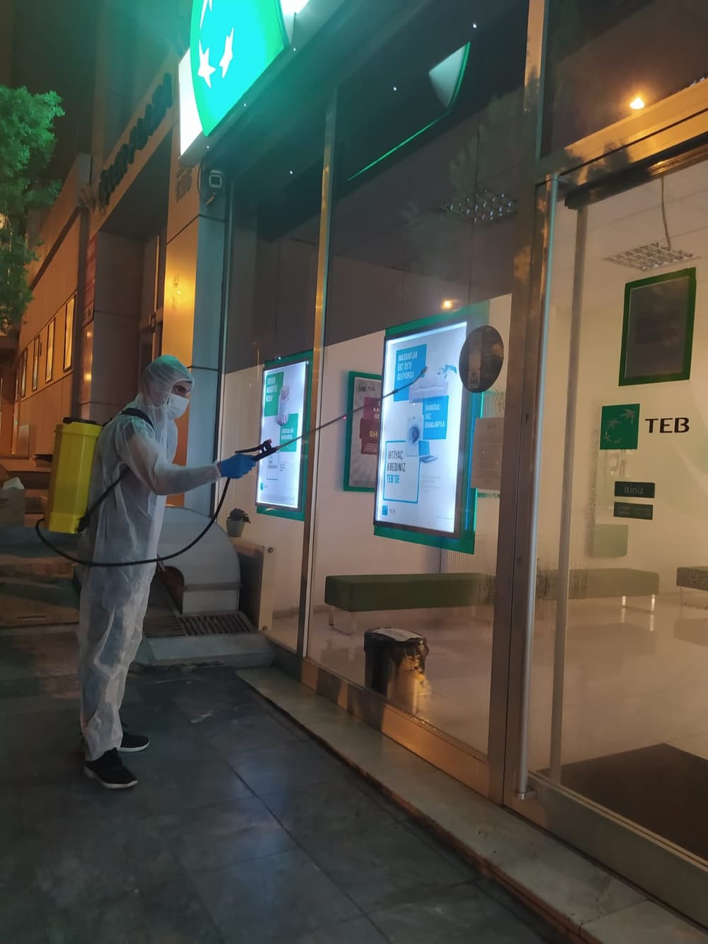 Tam koruyucu tulum, maske ve eldiven takan uygulayıcı, gece saatlerinde bir banka şubesinin dış cephesini sırt pompasıyla ilaçlıyor